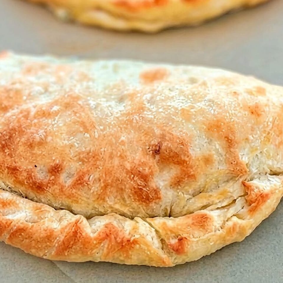 Golden-brown rustic stuffed pastry with a crimped edge rests on a baking sheet, with another pastry blurred in the background.