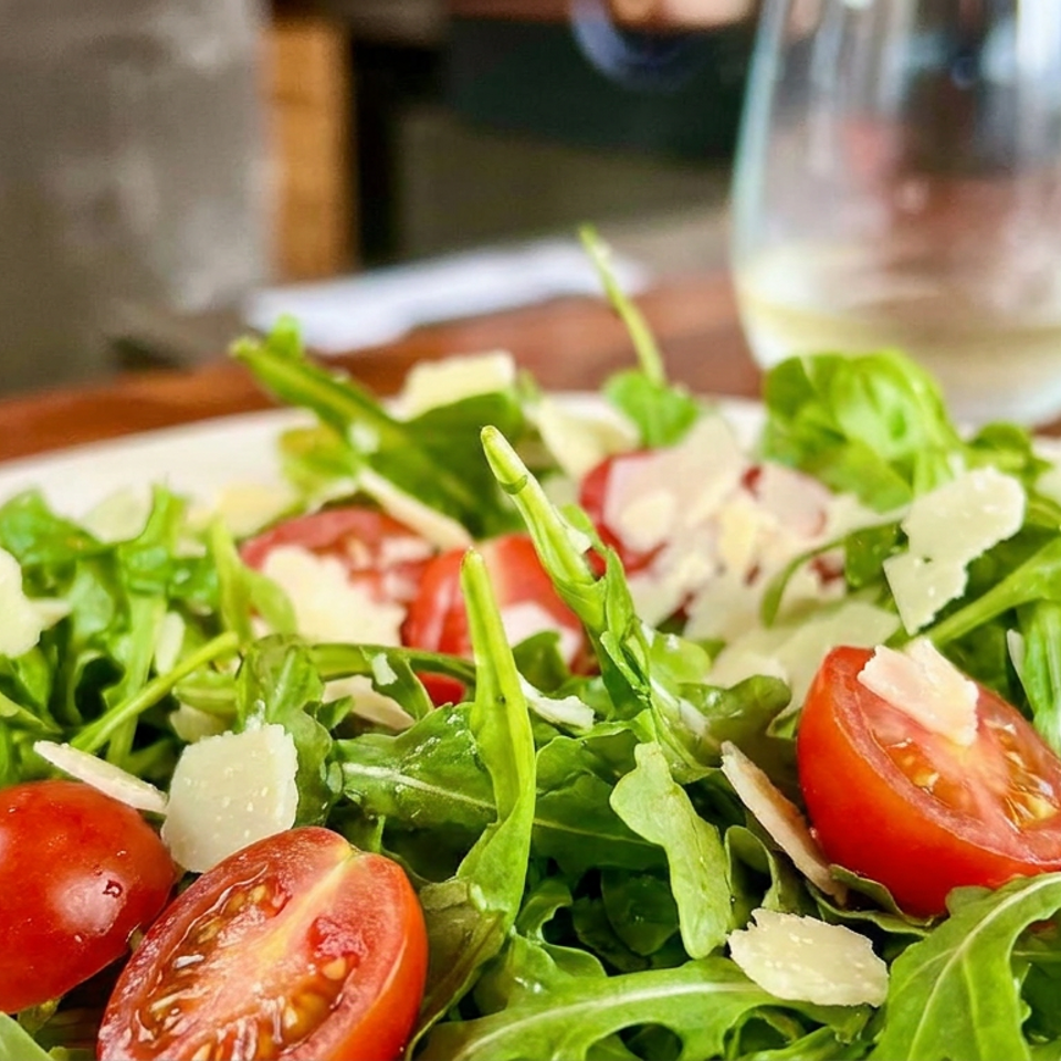 Fresh arugula salad with cherry tomatoes and shaved Parmesan served on a white plate beside a wine glass.
