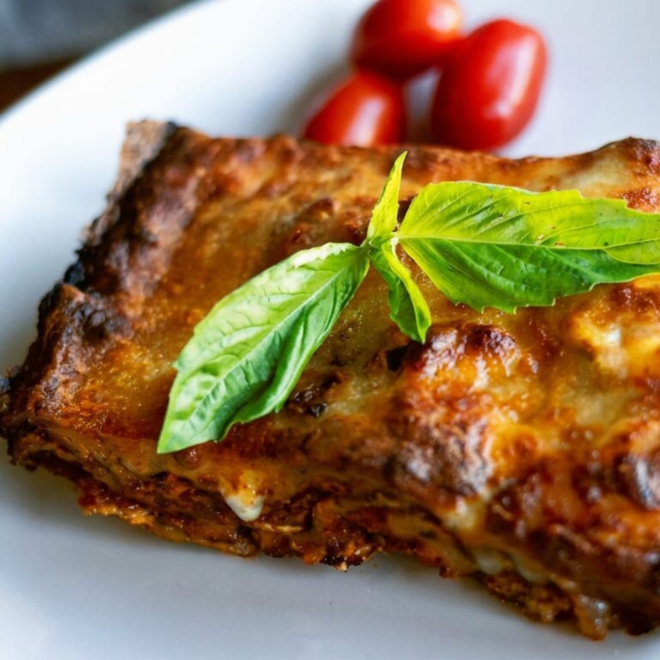 Golden-brown baked lasagna topped with fresh basil leaves and cherry tomatoes on a white plate.