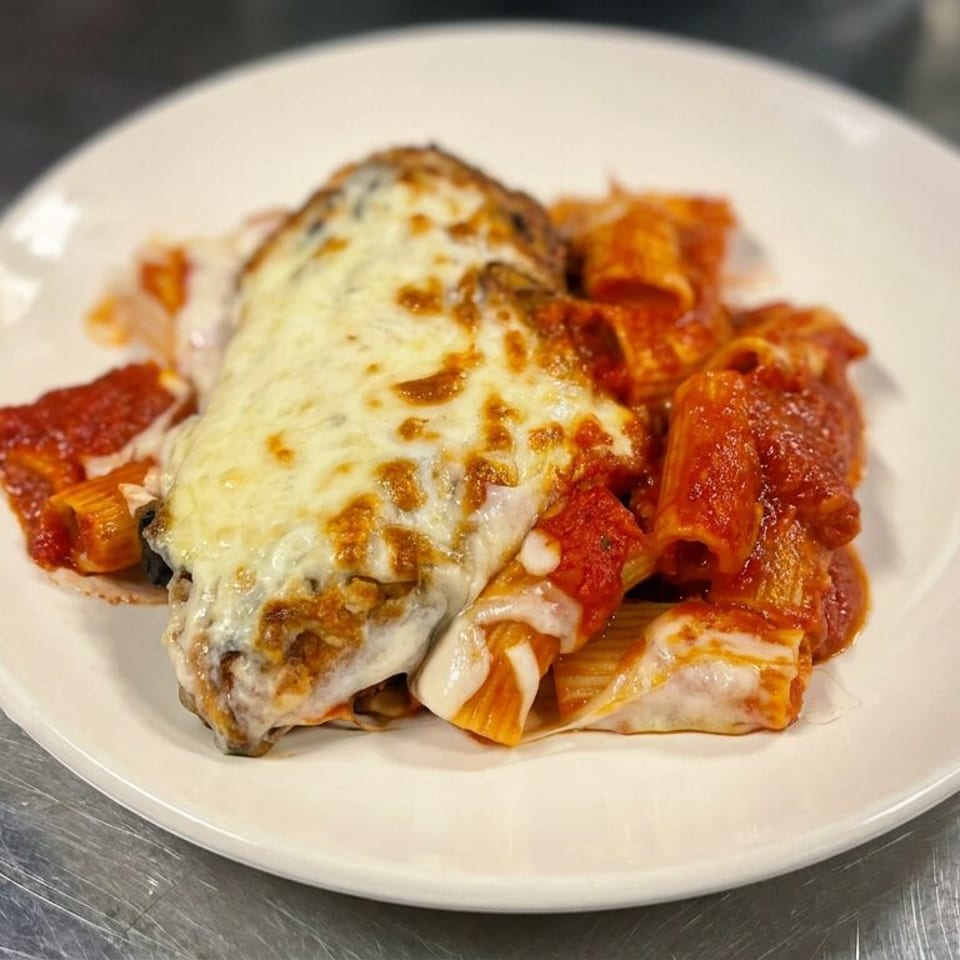Cheese-covered chicken breast served with tomato-sauced pasta on a white plate atop a stainless steel counter.