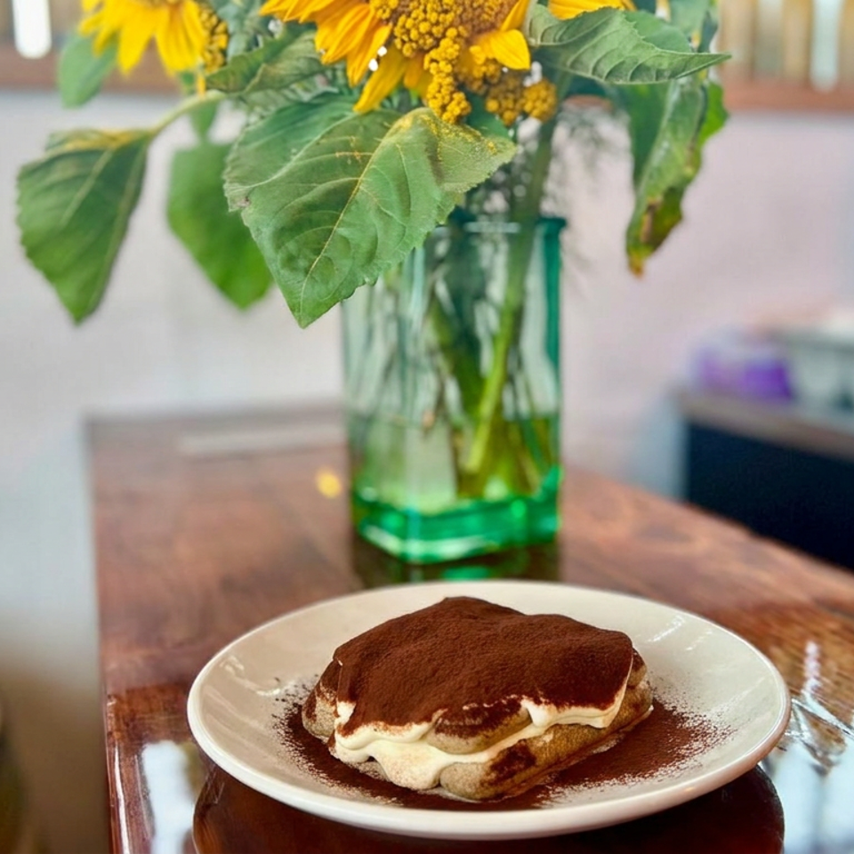 Tiramisu dusted with cocoa sits on a white plate atop a wooden restaurant table beside a sunflower-filled green glass vase.