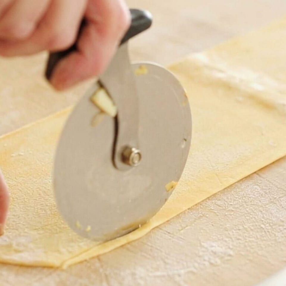 Hands use a circular cutter to slice fresh pasta dough on a floured work surface.